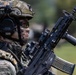 A heavily equipped military or special operations soldier is shown in a close-up profile view, wearing full tactical gear including a camouflage helmet, MSA hearing protection, and a face covering. The soldier is holding a modern assault rifle with visible accessories including an optic sight and what appears to be a loaded magazine. The background is blurred, suggesting an outdoor field or training environment.