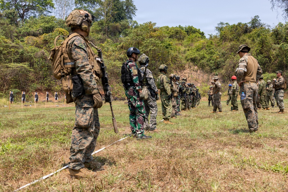 A multinational military training exercise is taking place on an outdoor firing range surrounded by dense vegetation. Multiple soldiers wearing different camouflage patterns and uniforms are lined up along a marked boundary line, suggesting a joint training event. Shooting targets are visible downrange in the background.