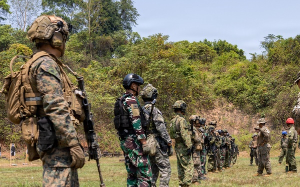 A multinational military training exercise is taking place on an outdoor firing range surrounded by dense vegetation. Multiple soldiers wearing different camouflage patterns and uniforms are lined up along a marked boundary line, suggesting a joint training event. Shooting targets are visible downrange in the background.