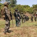 A multinational military training exercise is taking place on an outdoor firing range surrounded by dense vegetation. Multiple soldiers wearing different camouflage patterns and uniforms are lined up along a marked boundary line, suggesting a joint training event. Shooting targets are visible downrange in the background.