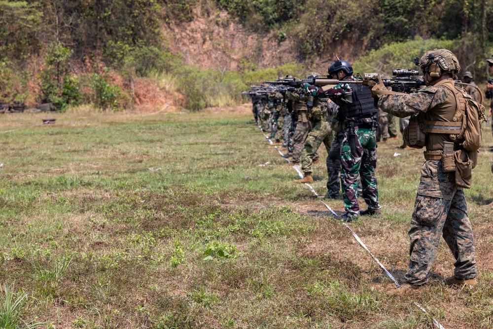 A line of military personnel are conducting a live-fire shooting exercise on an outdoor range. Two distinct uniform types are visible: woodland camouflage (consistent with Indonesian TNI pattern) and MARPAT digital camouflage (consistent with US Marine Corps). The soldiers are aligned along a firing line marked with white cord/rope, aiming rifles downrange toward a hillside backdrop.