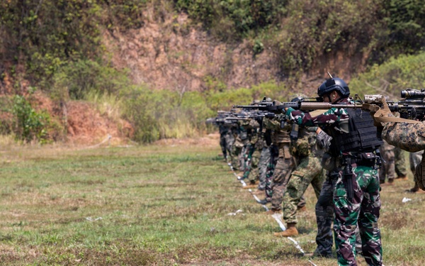 A line of military personnel are conducting a live-fire shooting exercise on an outdoor range. Two distinct uniform types are visible: woodland camouflage (consistent with Indonesian TNI pattern) and MARPAT digital camouflage (consistent with US Marine Corps). The soldiers are aligned along a firing line marked with white cord/rope, aiming rifles downrange toward a hillside backdrop.