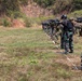 A line of military personnel are conducting a live-fire shooting exercise on an outdoor range. Two distinct uniform types are visible: woodland camouflage (consistent with Indonesian TNI pattern) and MARPAT digital camouflage (consistent with US Marine Corps). The soldiers are aligned along a firing line marked with white cord/rope, aiming rifles downrange toward a hillside backdrop.