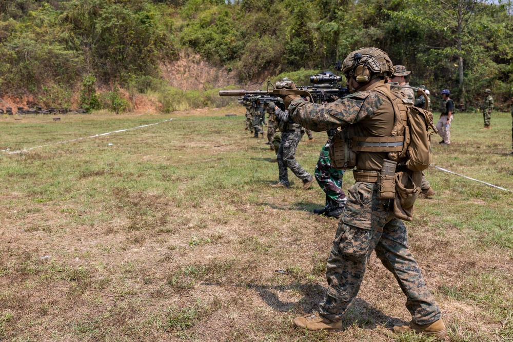 A military training exercise is taking place on an outdoor firing range, with multiple soldiers from what appear to be different nations lined up in a shooting formation. The foreground soldier wears USMC MARPAT digital camouflage with full tactical gear, while others in the line wear different camouflage patterns suggesting a multinational exercise. Observers and range safety personnel are visible in the background.