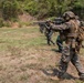 A military training exercise is taking place on an outdoor firing range, with multiple soldiers from what appear to be different nations lined up in a shooting formation. The foreground soldier wears USMC MARPAT digital camouflage with full tactical gear, while others in the line wear different camouflage patterns suggesting a multinational exercise. Observers and range safety personnel are visible in the background.