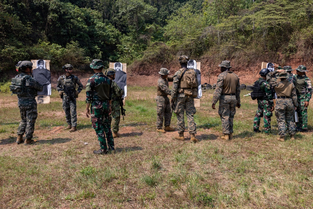 Military personnel from multiple nations are gathered at an outdoor shooting range conducting a joint training exercise. Soldiers in different camouflage uniforms are grouped around paper silhouette targets, with instructors appearing to demonstrate or debrief techniques. The setting is a cleared range area backed by a forested hillside with exposed red earth.