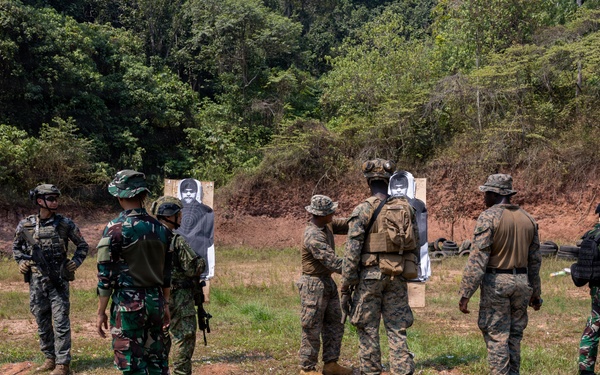 Military personnel from multiple nations are gathered at an outdoor shooting range conducting a joint training exercise. Soldiers in different camouflage uniforms are grouped around paper silhouette targets, with instructors appearing to demonstrate or debrief techniques. The setting is a cleared range area backed by a forested hillside with exposed red earth.