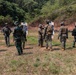 Military personnel from multiple nations are gathered at an outdoor shooting range conducting a joint training exercise. Soldiers in different camouflage uniforms are grouped around paper silhouette targets, with instructors appearing to demonstrate or debrief techniques. The setting is a cleared range area backed by a forested hillside with exposed red earth.