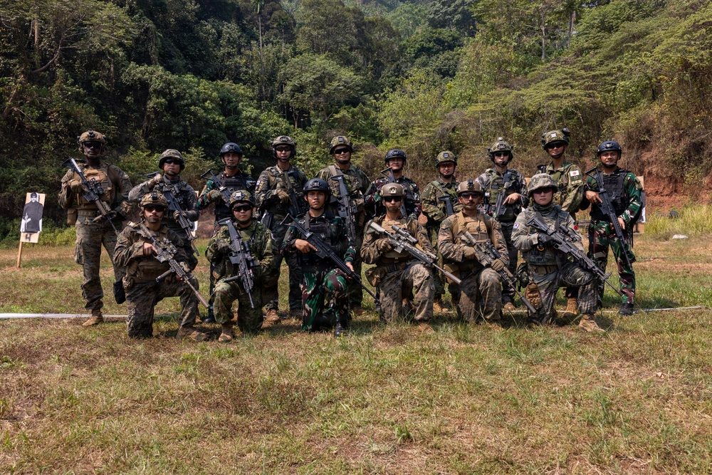 A group of approximately 16-17 armed military personnel pose for a group photograph at an outdoor shooting range surrounded by dense tropical forest. The soldiers appear to represent multiple nationalities based on their varying camouflage patterns and uniforms, suggesting a multinational joint military exercise or training event. Shooting targets are visible in the background to the left.