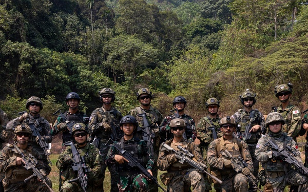 A group of approximately 16-17 armed military personnel pose for a group photograph at an outdoor shooting range surrounded by dense tropical forest. The soldiers appear to represent multiple nationalities based on their varying camouflage patterns and uniforms, suggesting a multinational joint military exercise or training event. Shooting targets are visible in the background to the left.