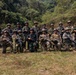 A group of approximately 16-17 armed military personnel pose for a group photograph at an outdoor shooting range surrounded by dense tropical forest. The soldiers appear to represent multiple nationalities based on their varying camouflage patterns and uniforms, suggesting a multinational joint military exercise or training event. Shooting targets are visible in the background to the left.