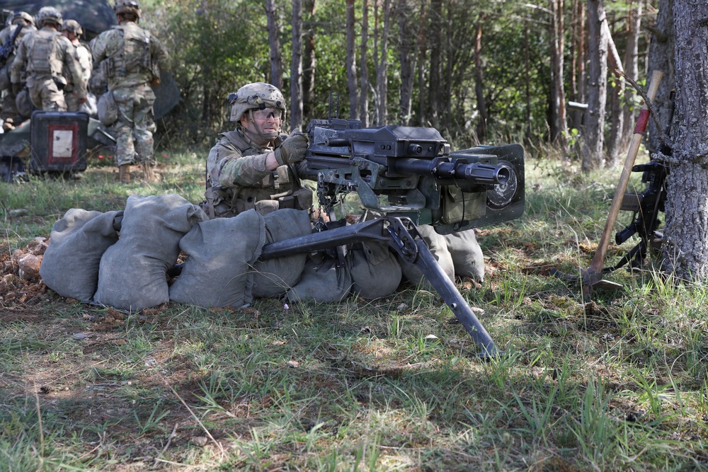 A soldier in full combat gear is positioned behind a sandbag emplacement operating what appears to be an M2 .50 caliber heavy machine gun or similar crew-served weapon mounted on a tripod. The setting is a wooded training area with additional soldiers visible in the background. The scene is consistent with a military field training exercise.