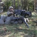 A soldier in full combat gear is positioned behind a sandbag emplacement operating what appears to be an M2 .50 caliber heavy machine gun or similar crew-served weapon mounted on a tripod. The setting is a wooded training area with additional soldiers visible in the background. The scene is consistent with a military field training exercise.