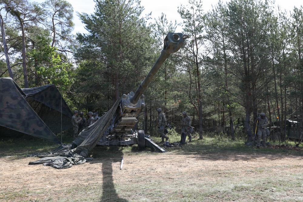 The image shows military personnel operating a large towed artillery piece in a forested clearing. Several soldiers in camouflage uniforms are positioned around the weapon system, which has its barrel elevated at a significant angle. Camouflage netting is deployed to the left side, suggesting a field position setup.