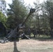 The image shows military personnel operating a large towed artillery piece in a forested clearing. Several soldiers in camouflage uniforms are positioned around the weapon system, which has its barrel elevated at a significant angle. Camouflage netting is deployed to the left side, suggesting a field position setup.