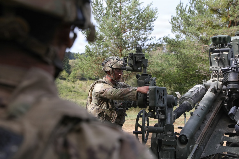 Two soldiers in modern U.S. Army MultiCam (OCP) camouflage uniforms and combat helmets are operating what appears to be an M777 155mm howitzer or similar towed artillery piece in an outdoor field setting. One soldier in the foreground is partially visible and out of focus, while a second soldier in the midground is actively adjusting the artillery piece's sighting or elevation mechanism. The setting appears to be a training area bordered by pine trees.