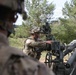 Two soldiers in modern U.S. Army MultiCam (OCP) camouflage uniforms and combat helmets are operating what appears to be an M777 155mm howitzer or similar towed artillery piece in an outdoor field setting. One soldier in the foreground is partially visible and out of focus, while a second soldier in the midground is actively adjusting the artillery piece's sighting or elevation mechanism. The setting appears to be a training area bordered by pine trees.