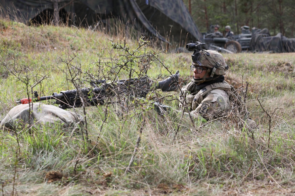 A soldier in full combat gear is in a prone firing position in a grassy field, partially concealed by sparse brush and vegetation. The soldier is equipped with a helmet-mounted device, glasses, and is operating what appears to be a machine gun with a red training blank-firing adapter attached to the muzzle. In the background, additional personnel and military vehicles are visible near a camouflage netting structure.