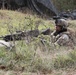 A soldier in full combat gear is in a prone firing position in a grassy field, partially concealed by sparse brush and vegetation. The soldier is equipped with a helmet-mounted device, glasses, and is operating what appears to be a machine gun with a red training blank-firing adapter attached to the muzzle. In the background, additional personnel and military vehicles are visible near a camouflage netting structure.