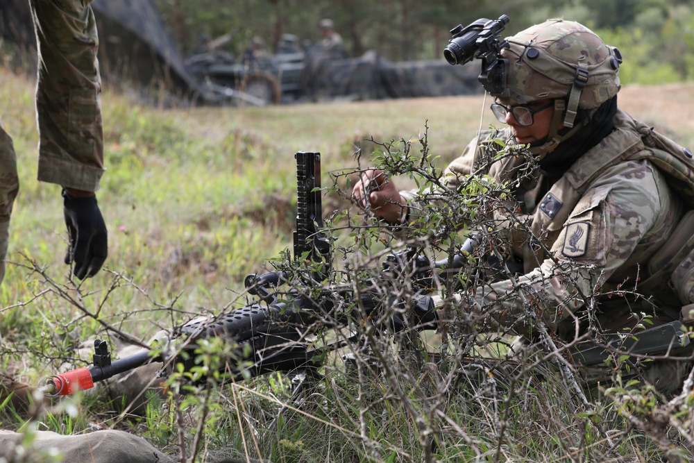 A soldier in full combat gear, including helmet with night vision mount and OCP (Operational Camouflage Pattern) uniform, is positioned low in a grassy field using natural brush for concealment. A weapon with a distinctive red training muzzle cap is visible in the foreground, indicating this is a training exercise rather than live combat. Additional personnel and military vehicles are visible in the background.