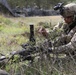A soldier in full combat gear, including helmet with night vision mount and OCP (Operational Camouflage Pattern) uniform, is positioned low in a grassy field using natural brush for concealment. A weapon with a distinctive red training muzzle cap is visible in the foreground, indicating this is a training exercise rather than live combat. Additional personnel and military vehicles are visible in the background.