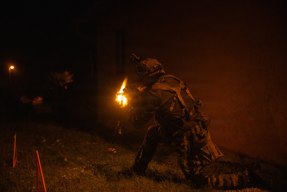 A heavily equipped soldier or tactical operator is photographed at night near a wall, with a visible muzzle flash or flame illuminating the scene. The individual is wearing full tactical gear including a helmet with mounted devices, body armor, and a loaded kit. The image is taken in low-light conditions with orange-tinted ambient smoke or haze visible.