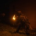 A heavily equipped soldier or tactical operator is photographed at night near a wall, with a visible muzzle flash or flame illuminating the scene. The individual is wearing full tactical gear including a helmet with mounted devices, body armor, and a loaded kit. The image is taken in low-light conditions with orange-tinted ambient smoke or haze visible.