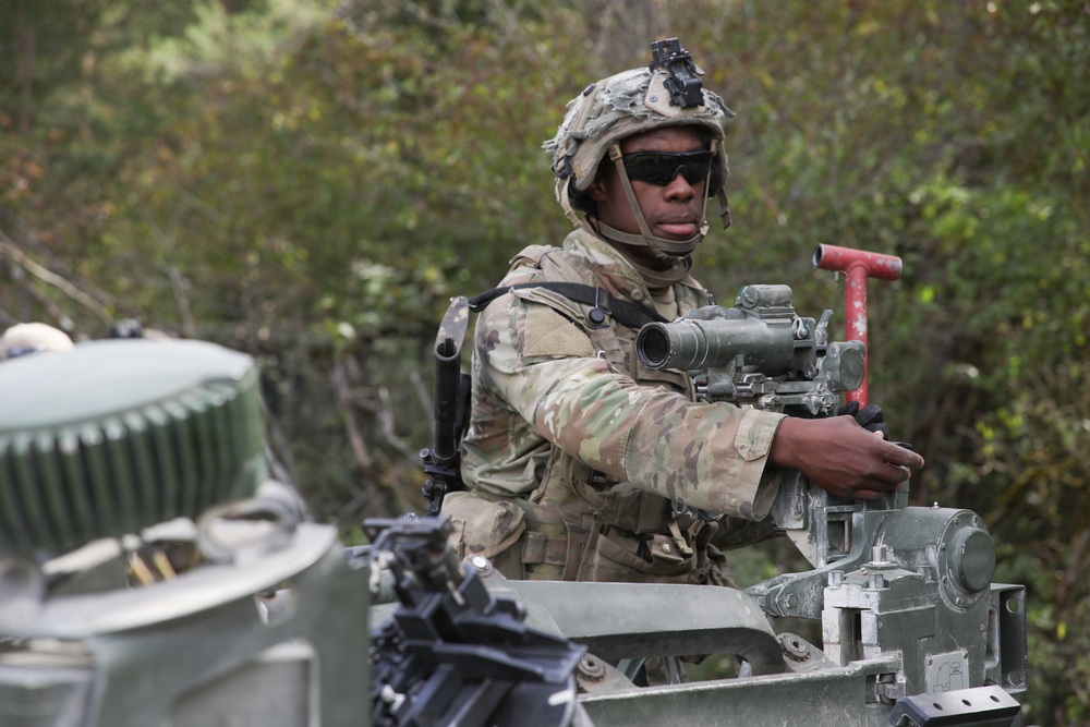 A soldier in full combat gear is positioned in the hatch or turret area of a military armored vehicle, appearing to operate or inspect a mounted weapon system. The soldier is wearing MultiCam (OCP) camouflage uniform, a combat helmet with night vision mount, and ballistic sunglasses. The setting appears to be a wooded or forested training or operational environment.