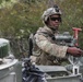 A soldier in full combat gear is positioned in the hatch or turret area of a military armored vehicle, appearing to operate or inspect a mounted weapon system. The soldier is wearing MultiCam (OCP) camouflage uniform, a combat helmet with night vision mount, and ballistic sunglasses. The setting appears to be a wooded or forested training or operational environment.