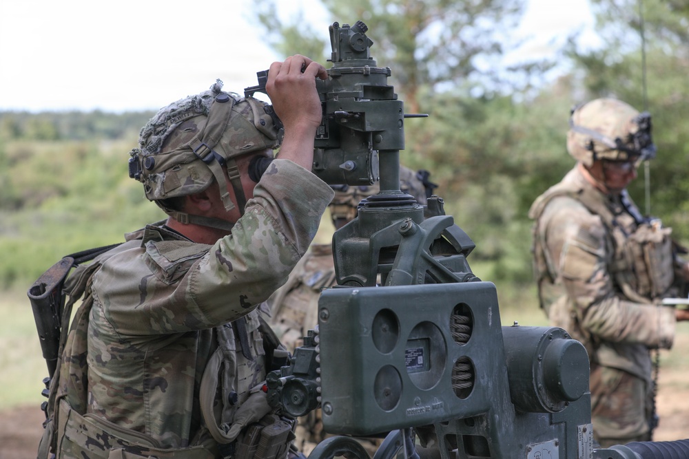 Two soldiers in full combat gear and camouflage uniforms are operating what appears to be a crew-served weapon system, likely an M2 .50 caliber machine gun or similar heavy weapon mounted on a tripod. The foreground soldier is looking through or adjusting the weapon's optical sight, while a second soldier stands in the background. The setting appears to be an outdoor training range or field exercise environment with trees visible in the background.