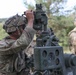 Two soldiers in full combat gear and camouflage uniforms are operating what appears to be a crew-served weapon system, likely an M2 .50 caliber machine gun or similar heavy weapon mounted on a tripod. The foreground soldier is looking through or adjusting the weapon's optical sight, while a second soldier stands in the background. The setting appears to be an outdoor training range or field exercise environment with trees visible in the background.