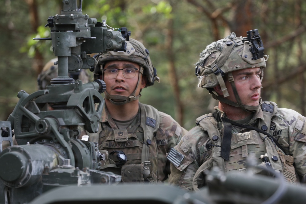 Two U.S. Army soldiers in full combat gear are positioned at what appears to be a military vehicle-mounted weapon system in a wooded environment. The soldier on the left, identified by name tape as 'ALVARADO,' wears glasses and a combat helmet with communications equipment. Both soldiers are wearing Operational Camouflage Pattern (OCP) uniforms with U.S. flag patches and appear to be on alert or conducting a training exercise.