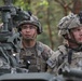 Two U.S. Army soldiers in full combat gear are positioned at what appears to be a military vehicle-mounted weapon system in a wooded environment. The soldier on the left, identified by name tape as 'ALVARADO,' wears glasses and a combat helmet with communications equipment. Both soldiers are wearing Operational Camouflage Pattern (OCP) uniforms with U.S. flag patches and appear to be on alert or conducting a training exercise.