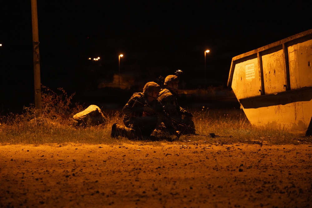 A nighttime scene showing two individuals in military-style camouflage gear taking a low prone or crouched position on open ground near a large industrial skip/dumpster. The scene is illuminated by warm orange ambient lighting, likely from nearby street lamps. The setting and posture suggest a military training exercise or tactical drill.