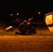 A nighttime scene showing two individuals in military-style camouflage gear taking a low prone or crouched position on open ground near a large industrial skip/dumpster. The scene is illuminated by warm orange ambient lighting, likely from nearby street lamps. The setting and posture suggest a military training exercise or tactical drill.