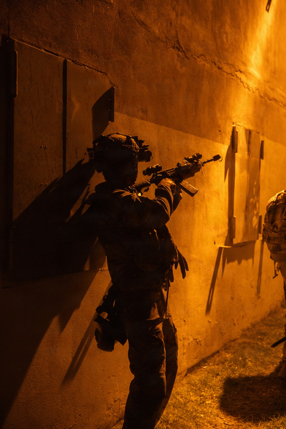 A heavily equipped tactical operator is moving along a wall in a narrow corridor or alleyway under intense orange ambient lighting, likely at night. The operator is armed and wearing night vision equipment on their helmet, suggesting a nighttime military or law enforcement operation. A second partially visible operator can be seen at the right edge of the frame.