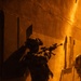 A heavily equipped tactical operator is moving along a wall in a narrow corridor or alleyway under intense orange ambient lighting, likely at night. The operator is armed and wearing night vision equipment on their helmet, suggesting a nighttime military or law enforcement operation. A second partially visible operator can be seen at the right edge of the frame.