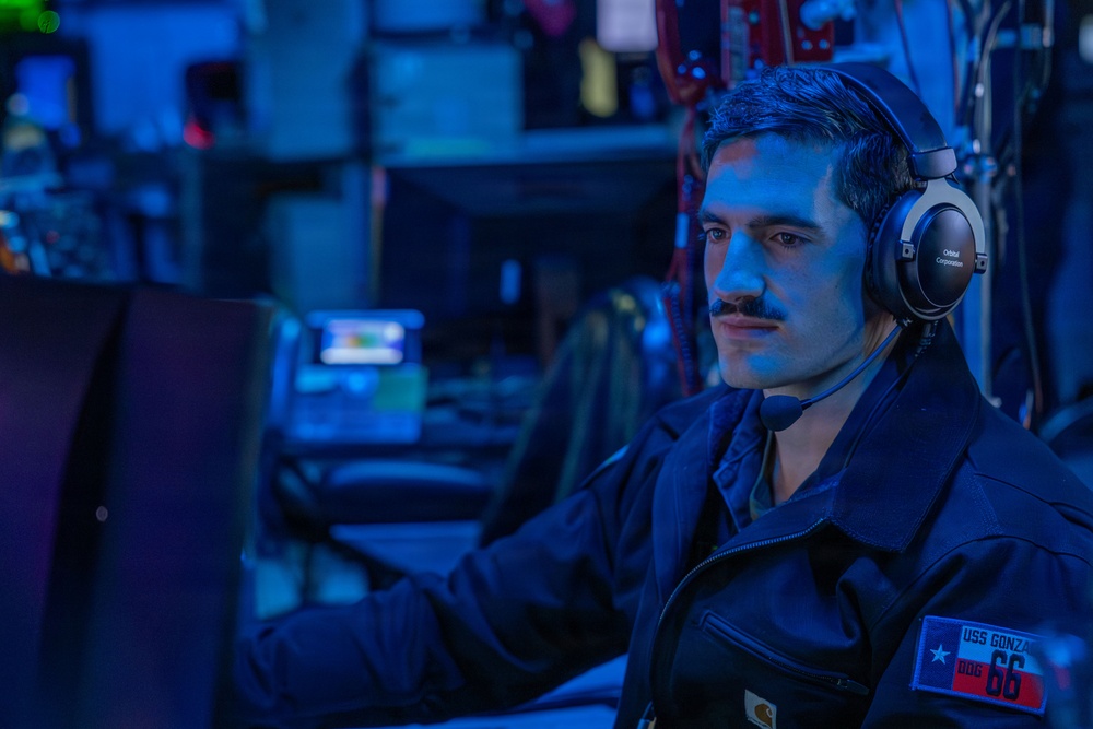 A male sailor wearing a dark navy uniform is seated at a workstation in what appears to be a combat information center or operations room aboard a naval vessel. He is wearing over-ear headphones labeled 'Orbital Corporation' and is focused on a monitor in front of him. The environment is illuminated with blue lighting, typical of shipboard operations centers.