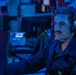 A male sailor wearing a dark navy uniform is seated at a workstation in what appears to be a combat information center or operations room aboard a naval vessel. He is wearing over-ear headphones labeled 'Orbital Corporation' and is focused on a monitor in front of him. The environment is illuminated with blue lighting, typical of shipboard operations centers.