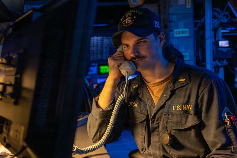 A U.S. Navy sailor identified as 'LUECK' is seated at a workstation aboard what appears to be a naval vessel, speaking on a corded telephone handset. He is wearing a dark blue USS Gonzalez ship's cap and a gray U.S. Navy coverall uniform. The scene is illuminated with a warm amber light contrasted against blue ambient lighting typical of a ship's combat information center or operations room.