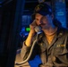 A U.S. Navy sailor identified as 'LUECK' is seated at a workstation aboard what appears to be a naval vessel, speaking on a corded telephone handset. He is wearing a dark blue USS Gonzalez ship's cap and a gray U.S. Navy coverall uniform. The scene is illuminated with a warm amber light contrasted against blue ambient lighting typical of a ship's combat information center or operations room.