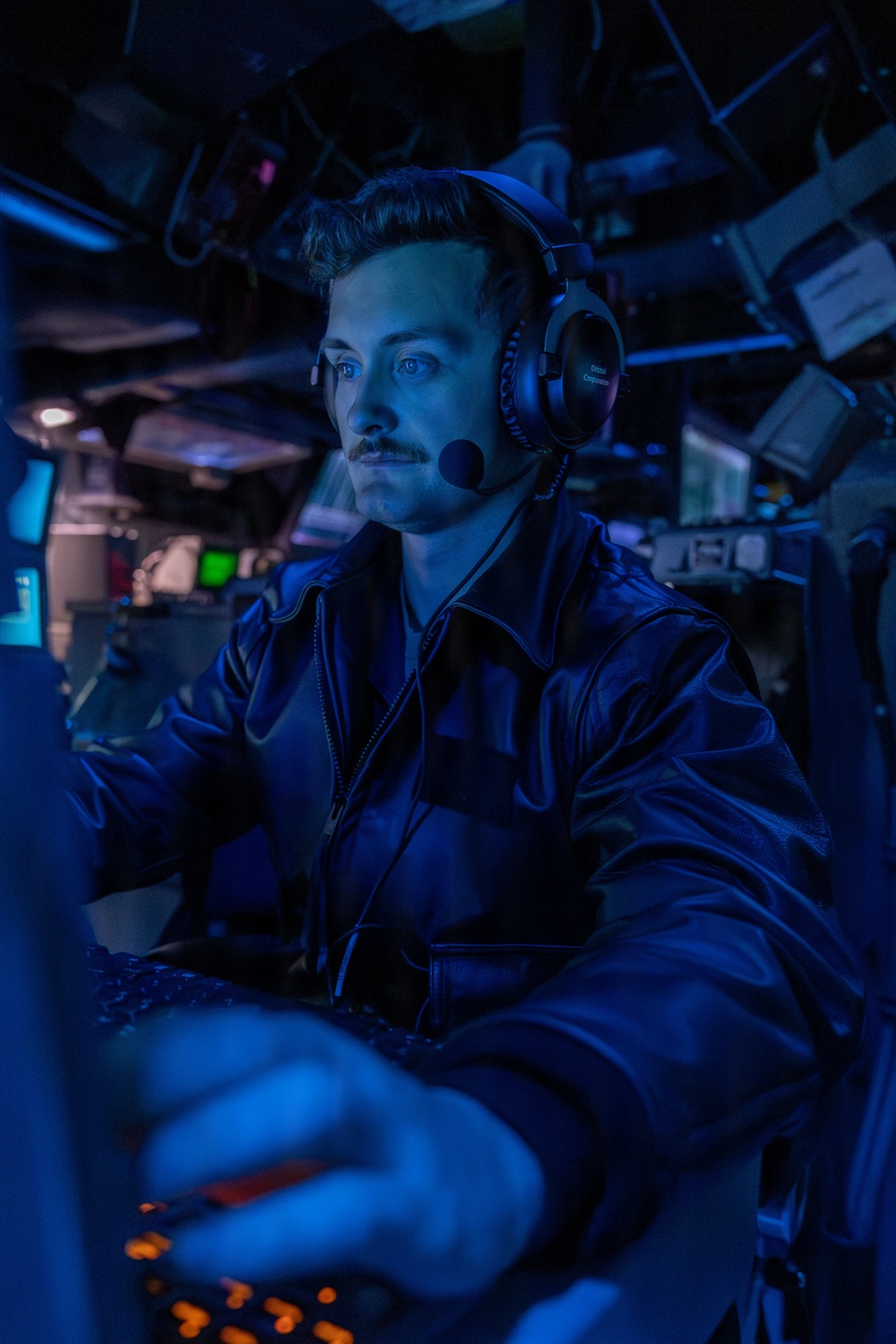 A young male operator wearing a headset with boom microphone is seated at a control station in a dimly lit, blue-illuminated combat information center or similar naval operations room. He is wearing a dark zip-up jacket and appears focused on equipment in front of him. Multiple screens and control panels are visible in the background.