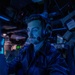 A young male operator wearing a headset with boom microphone is seated at a control station in a dimly lit, blue-illuminated combat information center or similar naval operations room. He is wearing a dark zip-up jacket and appears focused on equipment in front of him. Multiple screens and control panels are visible in the background.