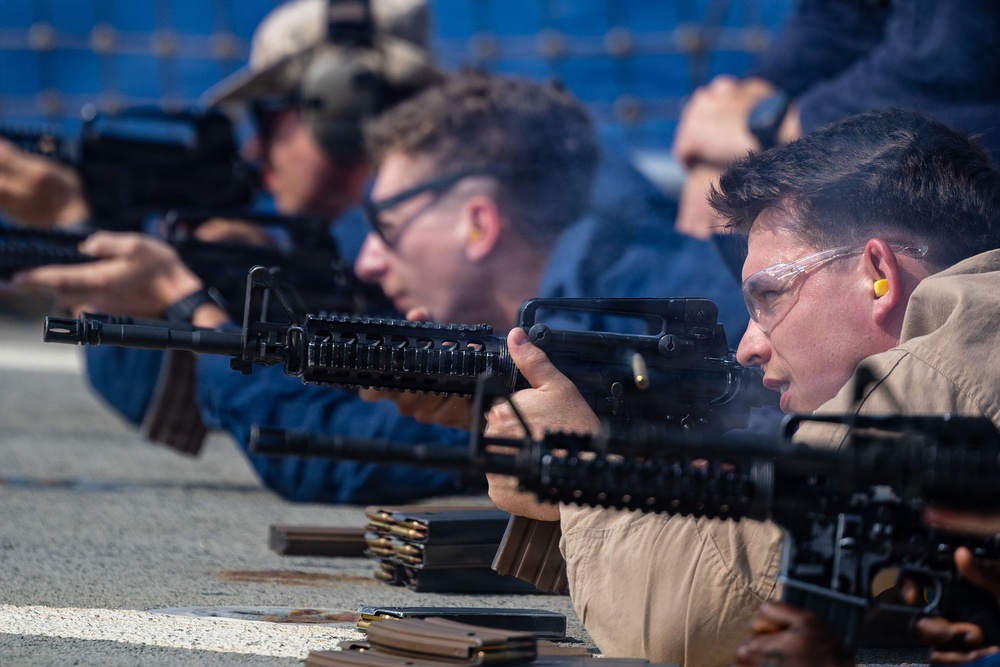 Multiple individuals are engaged in a live-fire shooting exercise at what appears to be a military or law enforcement range. They are lying in a prone or low position along a firing line, aiming M16/M4-style rifles downrange. Safety equipment including ear protection and protective eyewear is visible on the shooters.