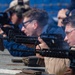 Multiple individuals are engaged in a live-fire shooting exercise at what appears to be a military or law enforcement range. They are lying in a prone or low position along a firing line, aiming M16/M4-style rifles downrange. Safety equipment including ear protection and protective eyewear is visible on the shooters.