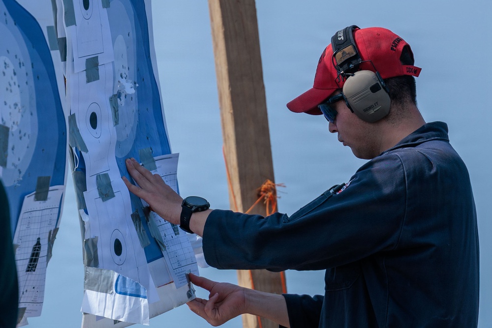 A man wearing a red baseball cap, hearing protection earmuffs, sunglasses, and a dark jacket is examining and patching a shooting target board. The target shows multiple concentric circle bullseye patterns with numerous bullet holes and gray patch stickers covering previous shots. The setting appears to be an outdoor shooting range near a body of water.