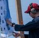 A man wearing a red baseball cap, hearing protection earmuffs, sunglasses, and a dark jacket is examining and patching a shooting target board. The target shows multiple concentric circle bullseye patterns with numerous bullet holes and gray patch stickers covering previous shots. The setting appears to be an outdoor shooting range near a body of water.
