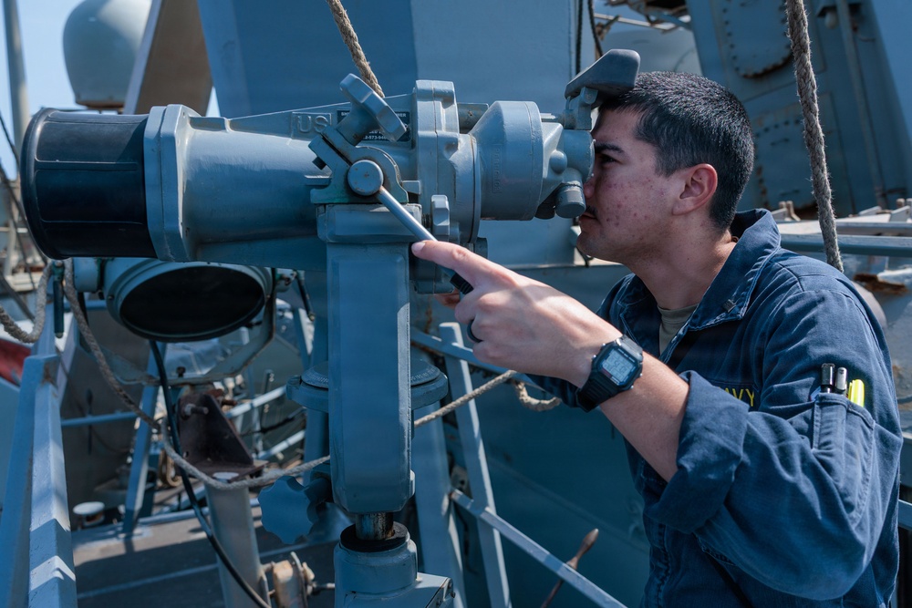 A U.S. Navy sailor in blue coveralls is looking through a large mounted optical instrument, likely a bearing circle or alidade, aboard a naval vessel. The sailor is pointing at an adjustment mechanism on the device while using it for observation or navigation. The setting is the exterior deck of a military ship with various naval equipment visible in the background.