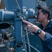 A U.S. Navy sailor in blue coveralls is looking through a large mounted optical instrument, likely a bearing circle or alidade, aboard a naval vessel. The sailor is pointing at an adjustment mechanism on the device while using it for observation or navigation. The setting is the exterior deck of a military ship with various naval equipment visible in the background.