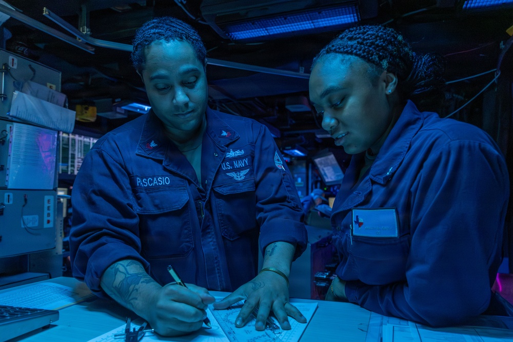 Two U.S. Navy personnel in blue utility uniforms are working together at a workstation aboard what appears to be a naval vessel. The individual on the left, identified by their name tape as 'PASCASIO,' is writing on a document while the other person observes. The environment is illuminated by blue lighting, typical of a ship's combat information center or navigation space.